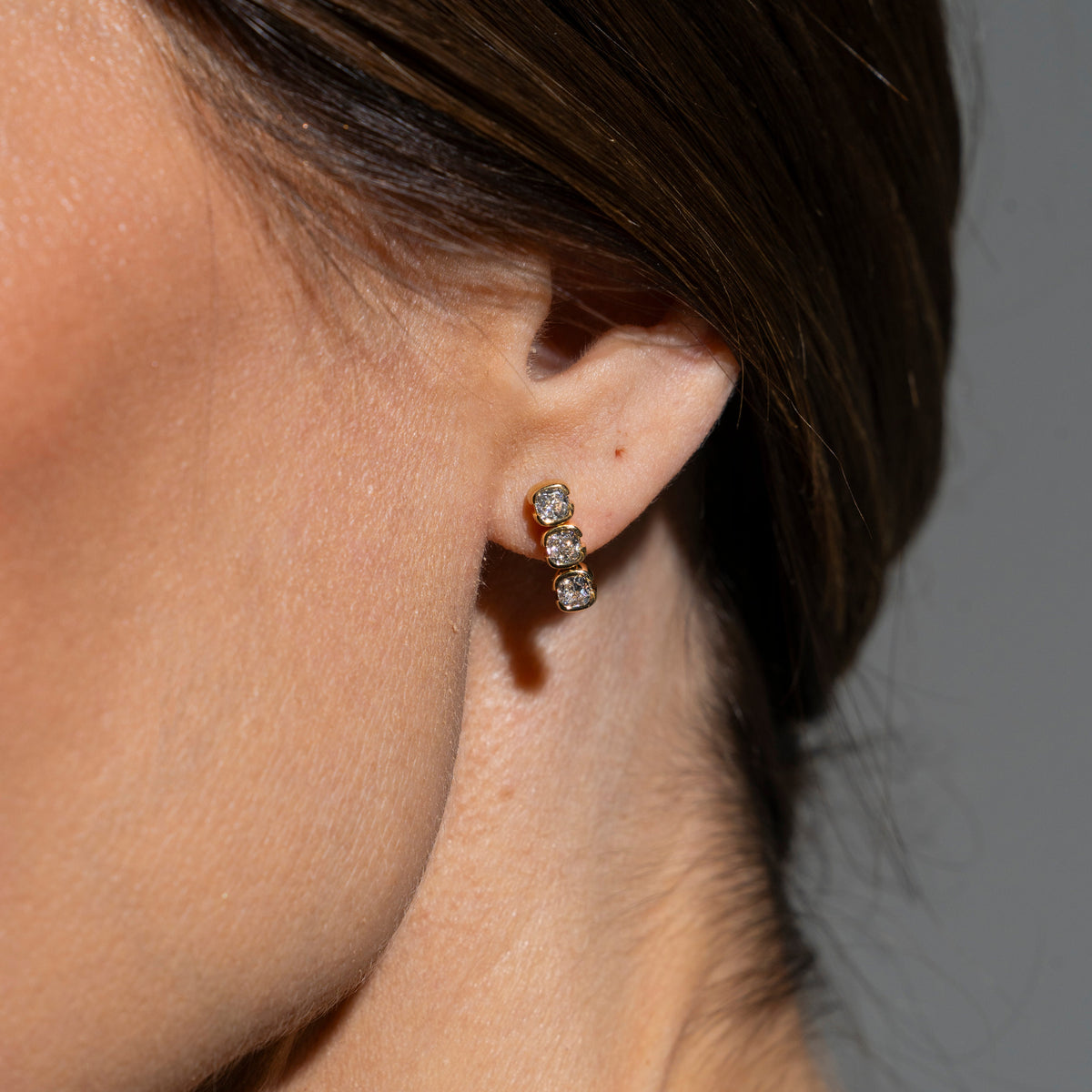 Close-up of an ear wearing a gold earring with three clear stones against a neutral background