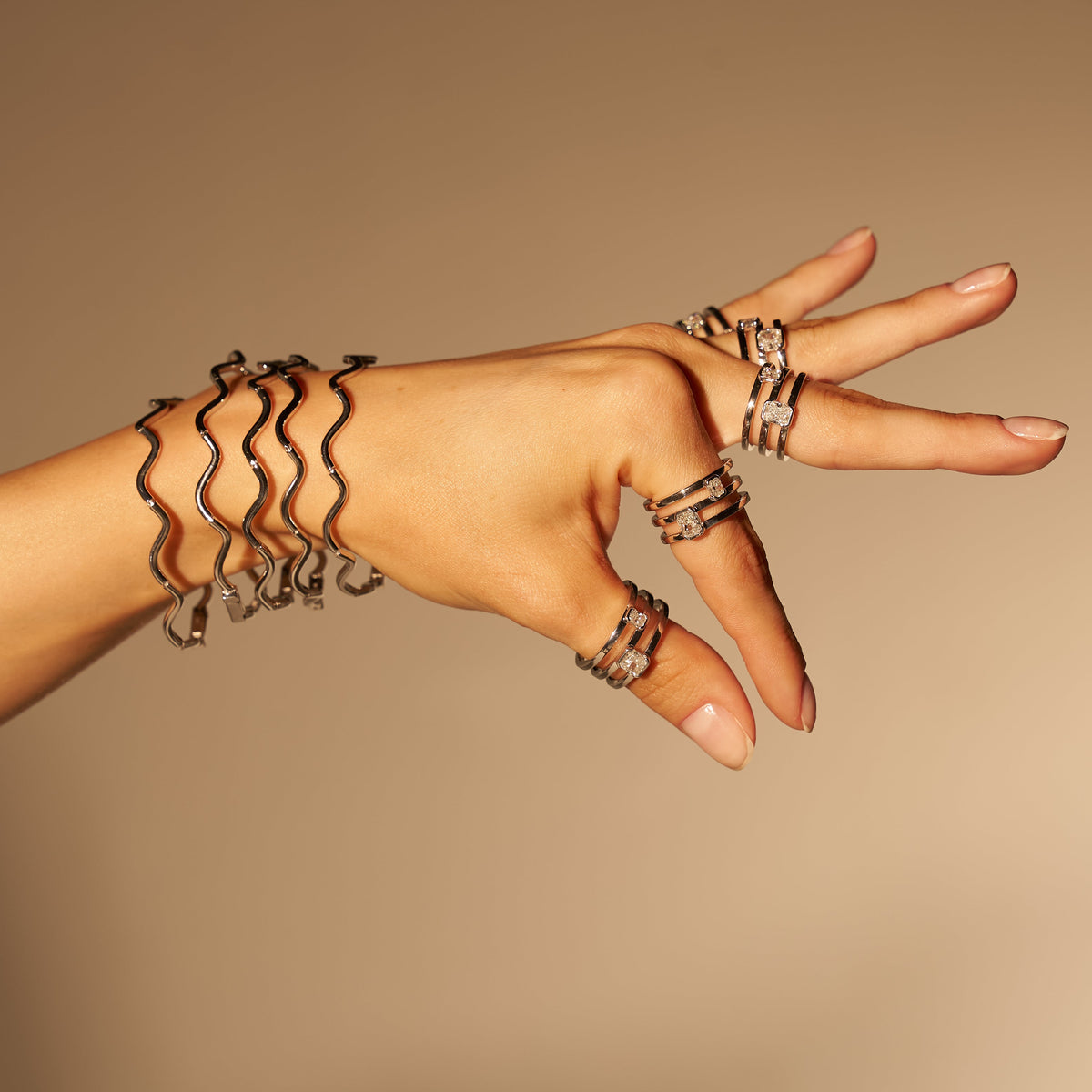 A hand wearing multiple silver wavy ring-style bracelets and matching stacked ring sets, posed against a smooth warm beige studio background.