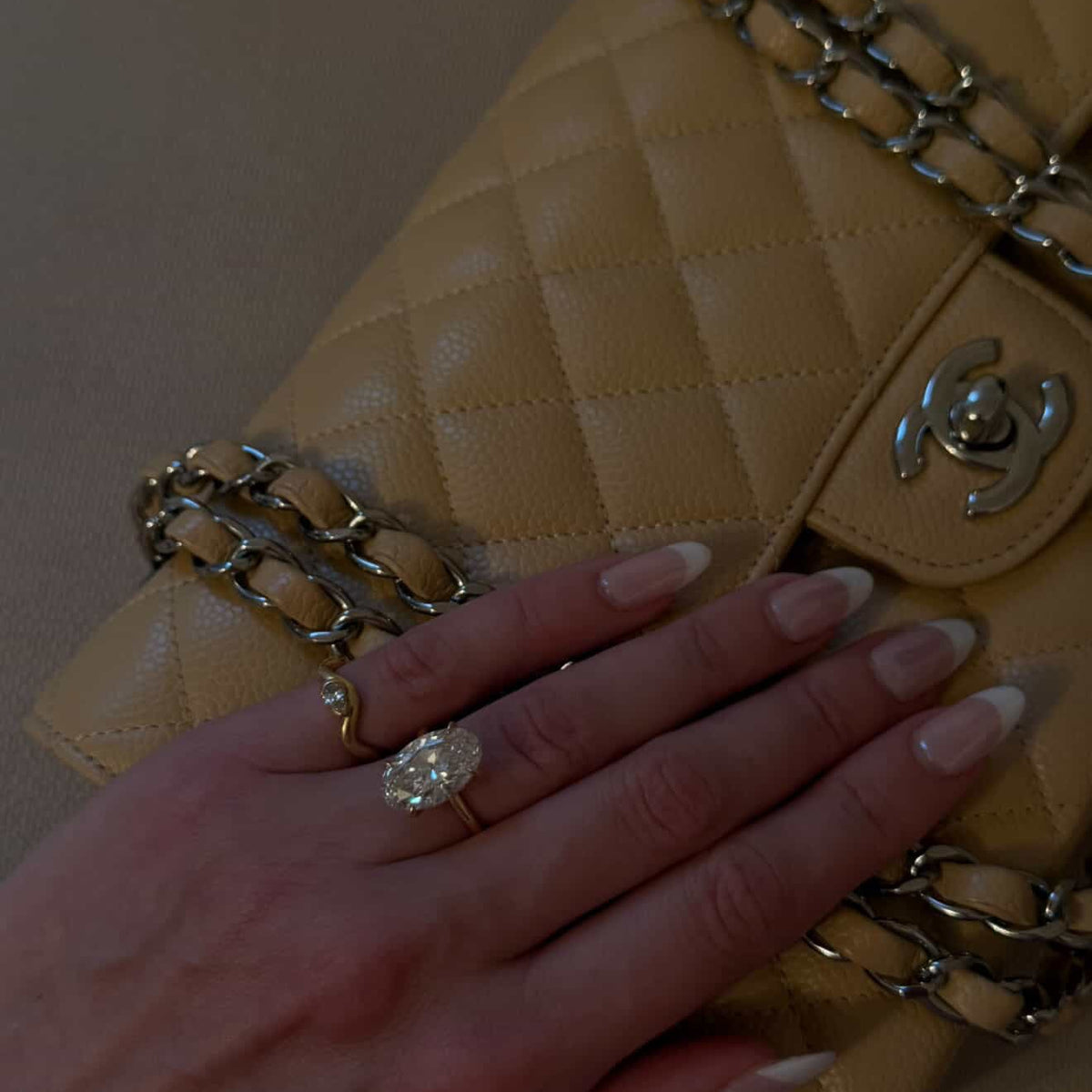 Woman wearing a dainty pinky ring next to her Oval Solitaire Engagement ring on top of her butter yellow chanel purse