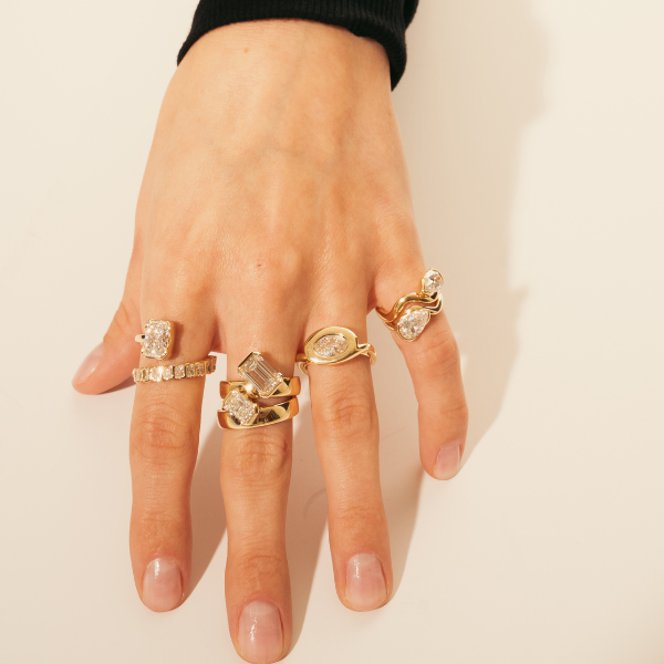 Hand wearing multiple gold rings with clear gemstones on a beige background
