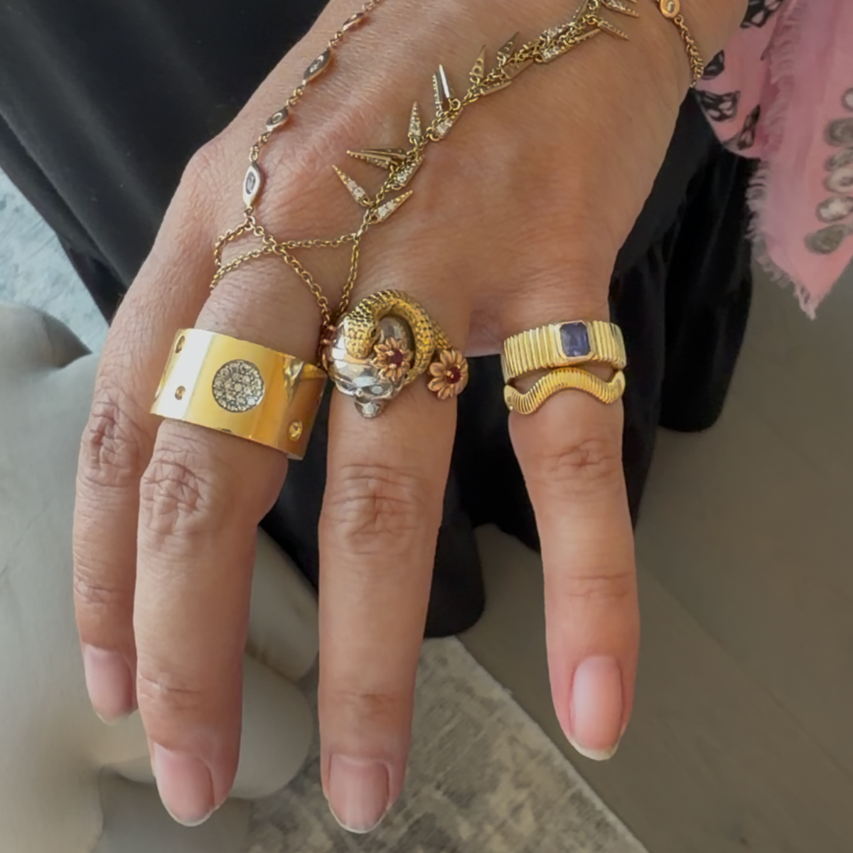 A close-up of a hand wearing multiple gold rings and a delicate gold chain bracelet with leaf charms. The rings include a wide yellow-gold band with a circular inset, a textured gold ring with a lion head motif, and a wavy gold band with a blue stone accent.