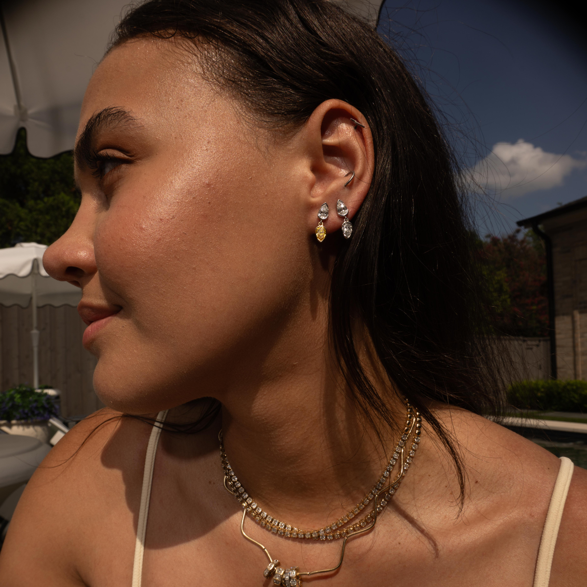 A close-up side profile of a woman wearing gold hoop earrings with clear stones and a small yellow accent, layered with a beaded gold necklace and a chain necklace, in an outdoor setting.