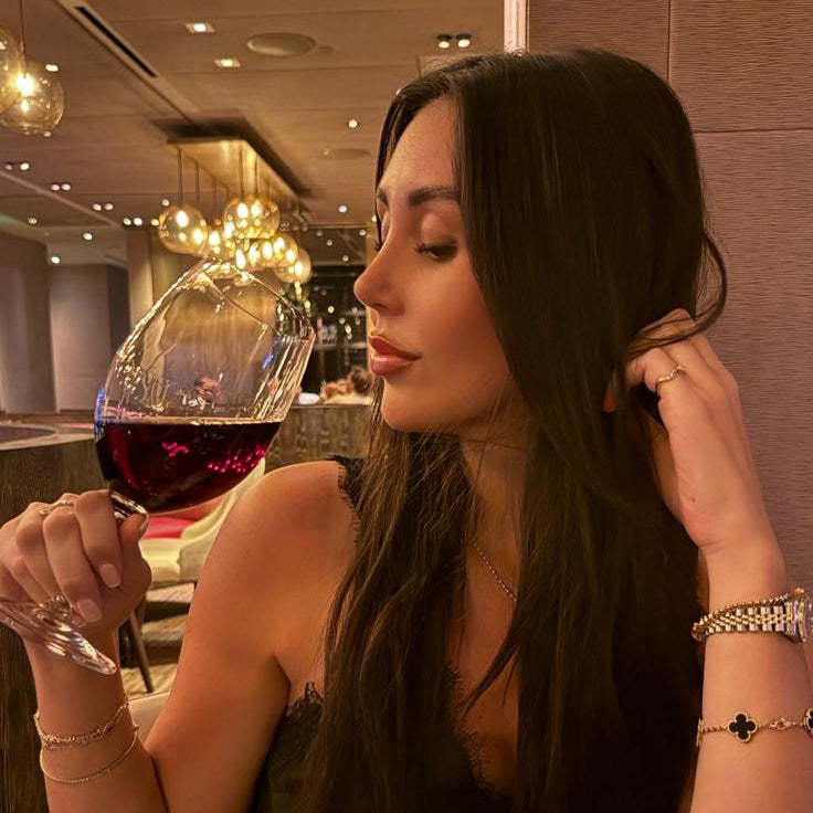 A woman with long dark hair sits in a restaurant setting, holding a glass of red wine. She is wearing a black sleeveless top and multiple silver-toned bracelets, with warm ambient lighting from chandeliers in the background.