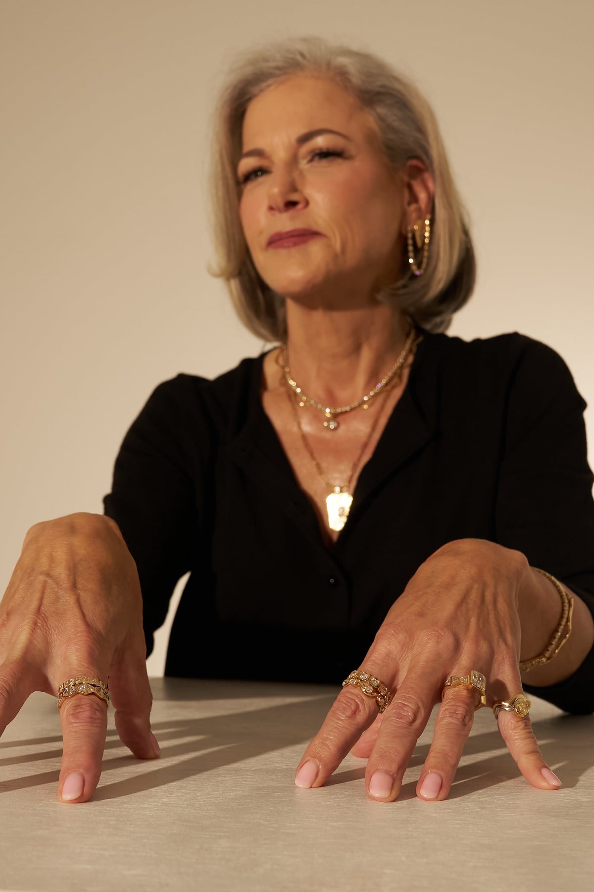 An older woman with short gray hair sits behind a table, wearing a black top and multiple gold rings and necklaces. Her hands are resting on the tabletop against a plain beige studio background.
