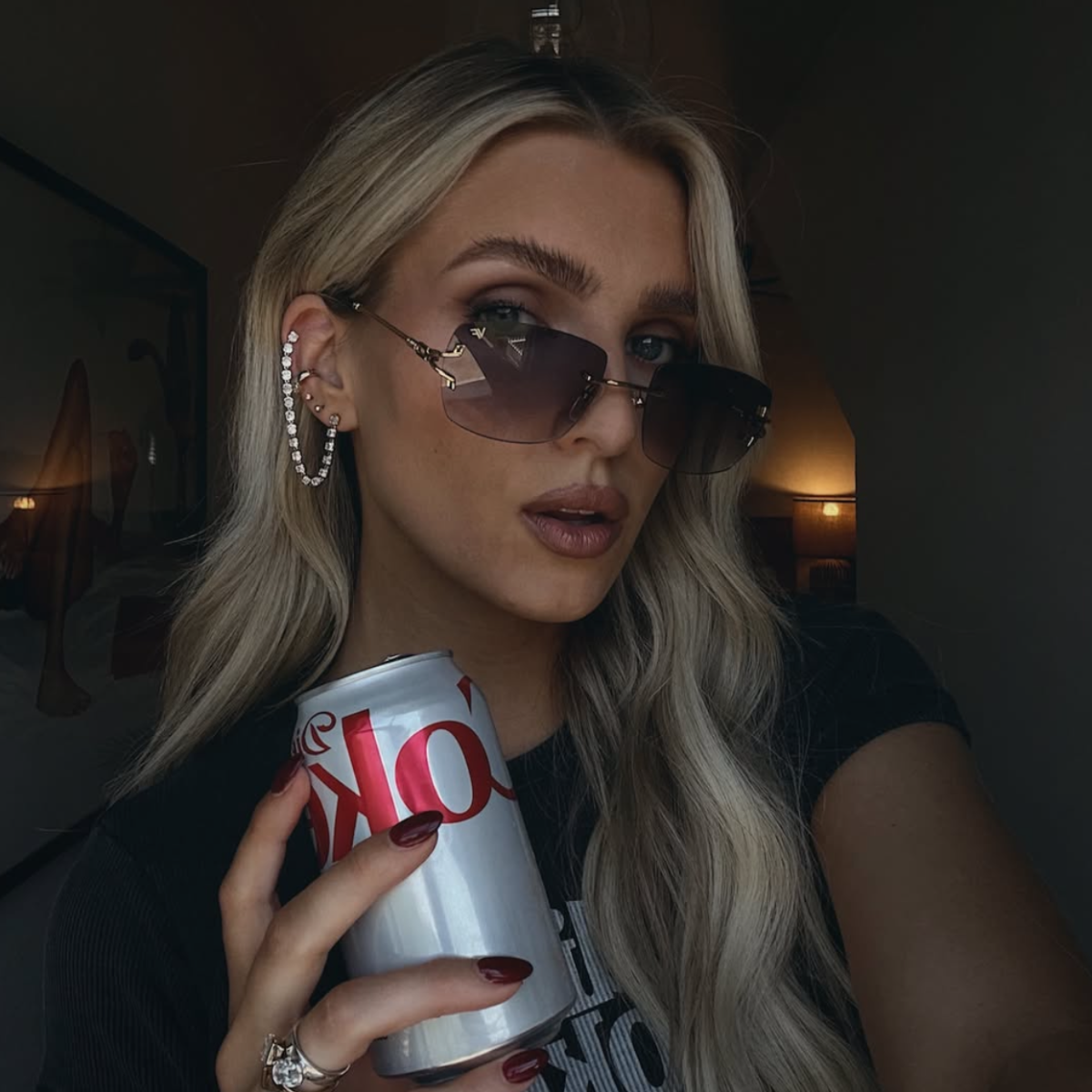 A woman with long blonde hair wearing gold-framed rimless sunglasses and holding a silver can of Coca-Cola, photographed in a dimly lit indoor setting.