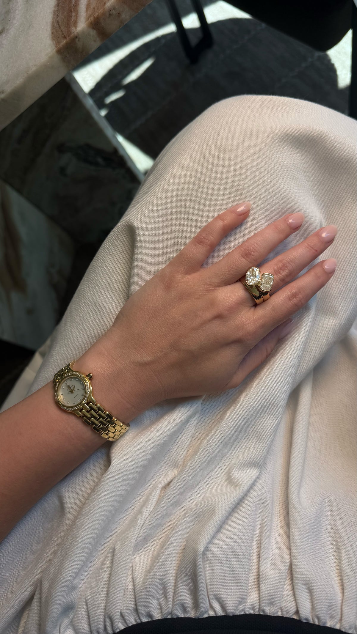 A close-up of a person's hand resting on a cream-colored fabric surface, wearing a gold-tone watch on the wrist and a large gold ring with a heart-shaped clear gemstone on the ring finger.