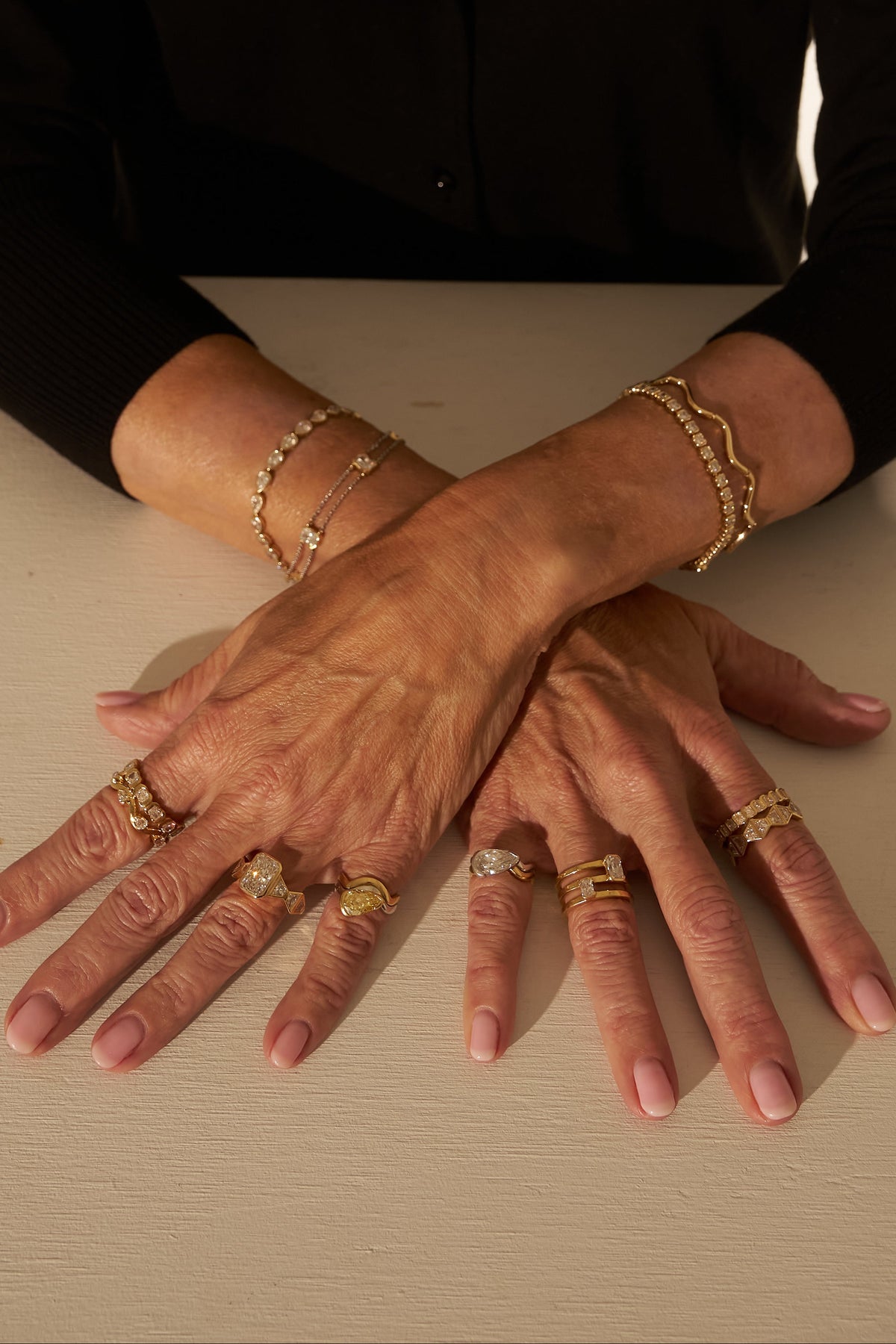 A close-up of a person's hands resting on a light beige table, adorned with multiple gold rings and bracelets, wearing a black cardigan and layered gold necklaces against a neutral beige background.